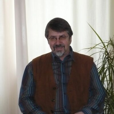 Ein Bild eines Mannes vor grauem Hintergrund. Er trägt ein gestreiftes Hemd und darüber eine braune Weste.